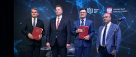 Czterech mężczyzn w garniturach stoi na tle granatowej ściany, dwóch trzyma czerwone teczki.