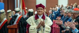 Mężczyzna w czerwonym ceremonialnym stroju z gronostajowym kołnierzem stoi i bije brawo, za nim wypełniona ludźmi sala.