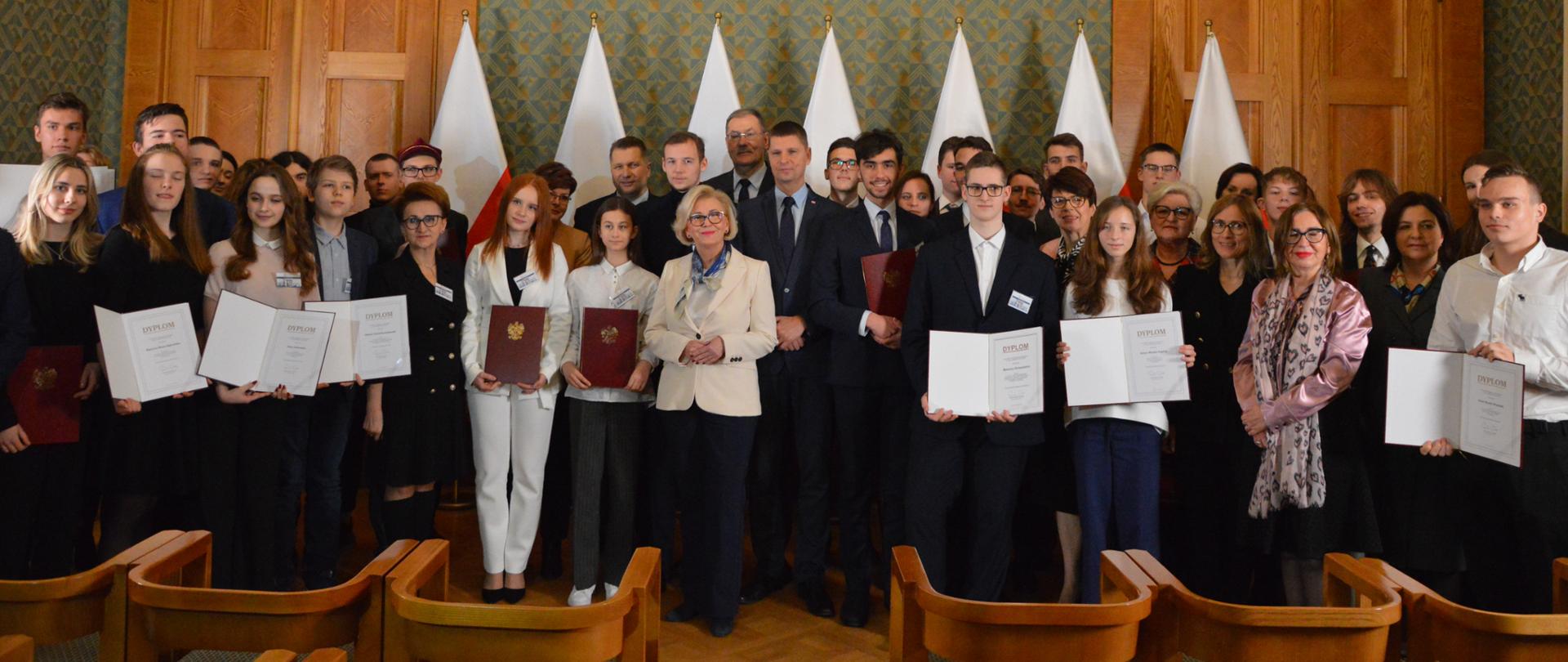 Przed zieloną wyłożoną drewnianymi panelami ścianą stoi duża grupa ludzi, niektórzy trzymają teczki, za nimi rząd siedmiu flag.