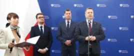 Na tle niebieskiej ściany stoi trzech mężczyzn w garniturach i kobieta w białej marynarce, trzymająca w ręku bezprzewodowy mikrofon. Za nimi z boku polska flaga.