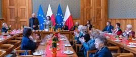 Widok na salę konferencyjną z długimi stołami nakrytymi czerwonymi bieżnikami. Na środku, pod flagami Polski i Unii Europejskiej, stoją przemawiające osoby. Seniorzy siedzący przy stołach klaszczą.