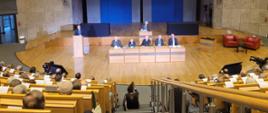 Widok z góry na dużą salę, na środku stół za którym siedzi pięciu prelegentów, za nimi na ścianie wiszą dwa wielkie ekrany na których widać kogoś mówiącego do mikrofonu.