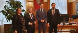Wiceminister Piontkowski, ubrana na brązowo kobieta i dwóch mężczyzn w garniturach stoją w eleganckim pokoju, za nimi drewniany stolik, duża roślina w doniczce, dalej żółta ściana pokryta drewnianymi panelami na której wisi godło Polski i dwa okna.