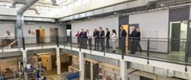 Widok na kilkupiętrową halę, na galeryjce stoi grupa osób, pod sufitem i przy ścianach biegną grube metalowe rury, na podłodze stoją różne urządzenia.