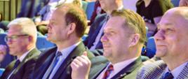Zdjęcie z boku na widownię, na ustawionych w rzędy krzesłach siedzą ludzie, z przodu minister Czarnek w otoczeniu kilku mężczyzn w garniturach.
