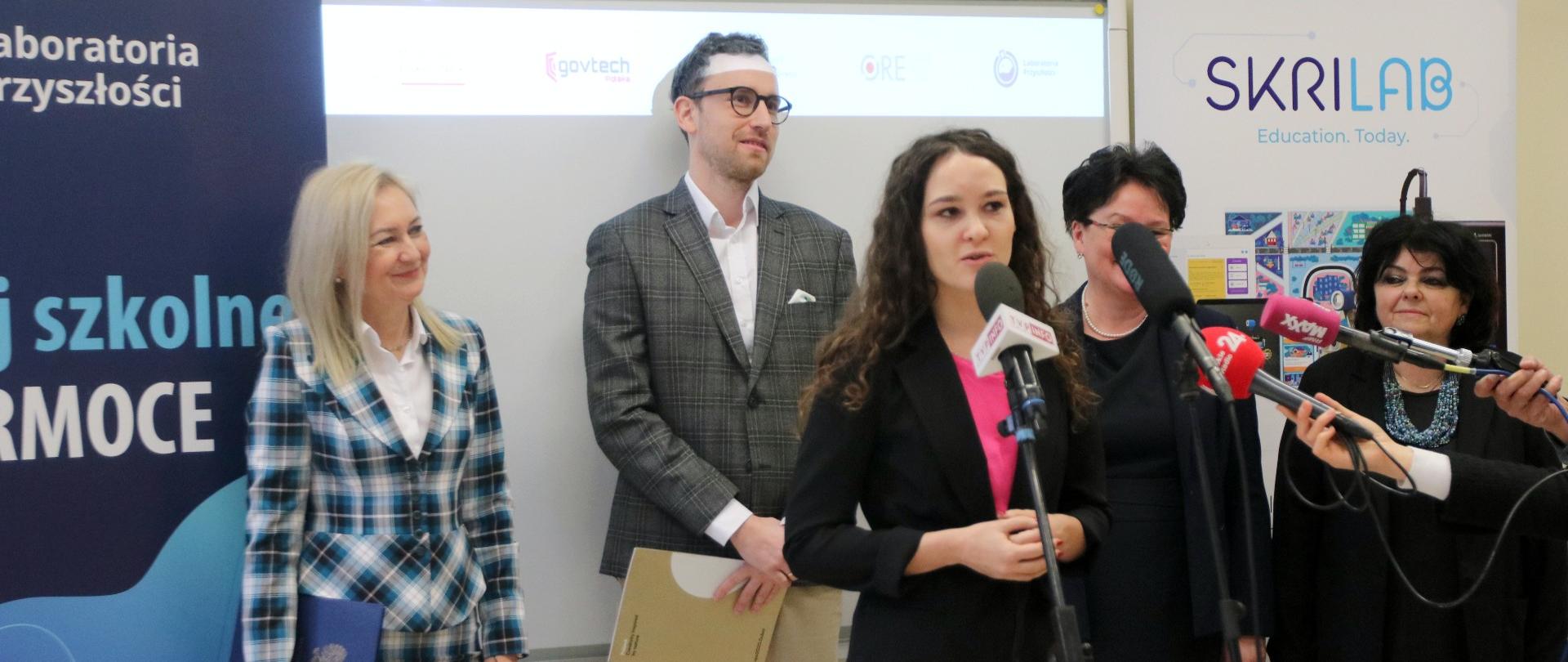 Ubrana na czarno kobieta mówi do mikrofonu na stojaku, za nią stoi kilka osób.