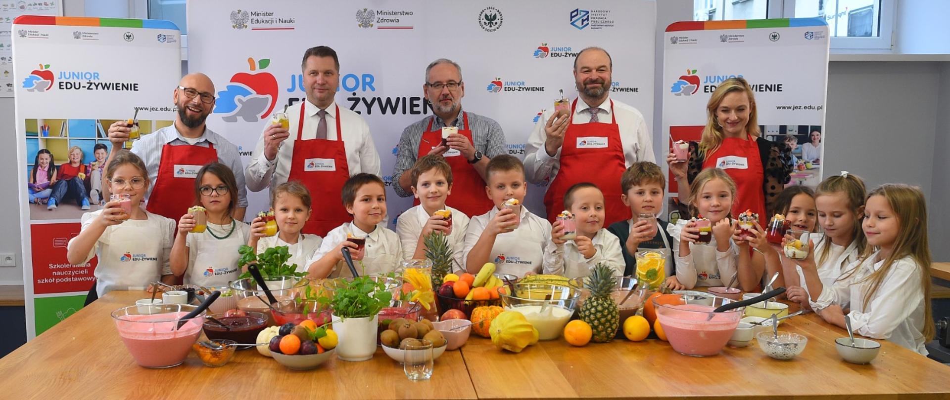 Grupa małych dzieci w białych fartuszkach stoi za stołem, na którym jest dużo kolorowych owoców, jedzą coś z naczyń trzymanych w rękach, za nimi stoi pięcioro dorosłych w czerwonych fartuchach, dalej ścianka z napisem Junior Edu-żywienie.