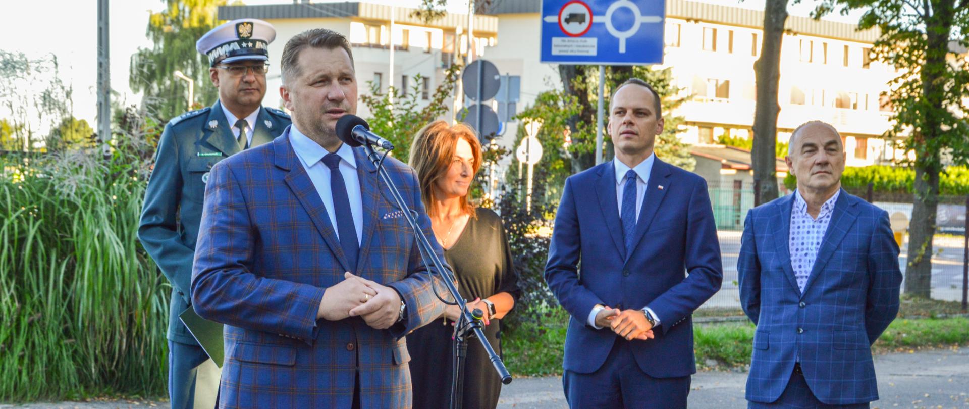 Minister Czarnek stoi przy mikrofonie. Za nim mężczyzna ubrany w mundur Inspekcji Transportu Drogowego, obok kobieta ubrana w czarną sukienkę oraz dwóch mężczyzn w garniturach. W tle zielone zarośla i oświetlone porannym słońcem bloki mieszkalne. 