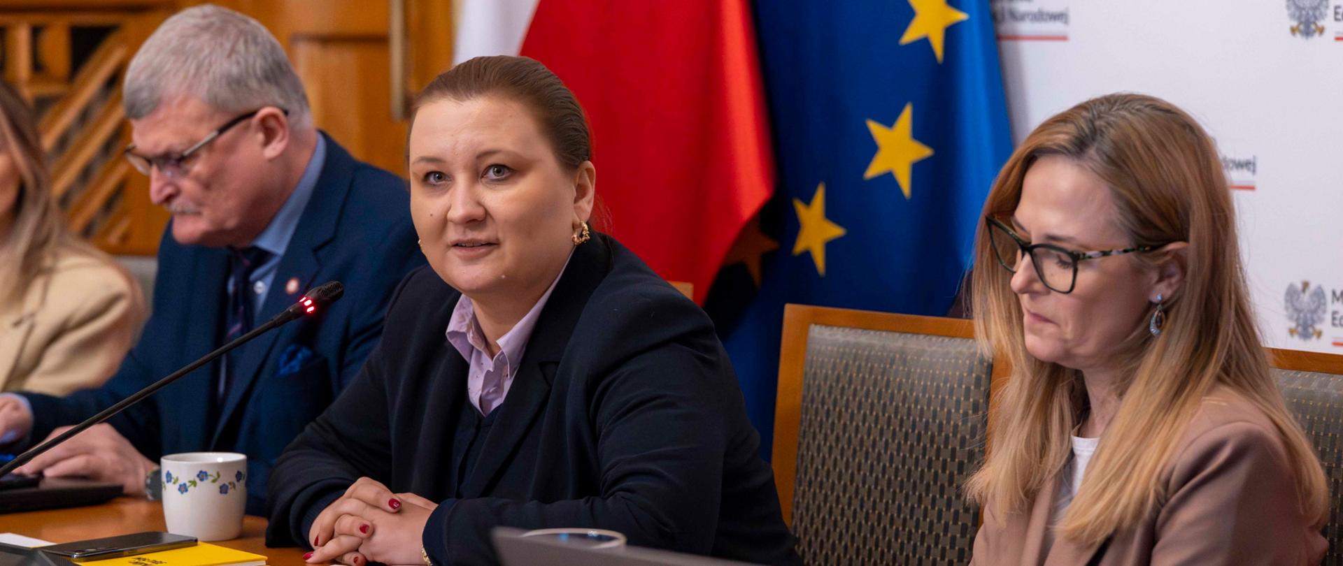 Zdjęcie przedstawia minister Paulinę Piechnę-Więckiewicz, podsekretarz stanu w Ministerstwie Edukacji Narodowej, która siedzi przy stole i przemawia do mikrofonu. Przed nią leżą materiały konferencyjne, telefon i kubek. Obok niej siedzą minister Katarzyna Kącka, podsekretarz stanu w Ministerstwie Zdrowia, a także Paweł Grzesiowski – Główny Inspektor Sanitarny. Postacie siedzą na tle flag Polski i Unii Europejskiej oraz ścianki z logotypami Ministerstwa Edukacji Narodowej. Kąt zdjęcia wskazuje na wykonywanie go pod kątem około 45 stopni względem postaci widocznych na fotografii.