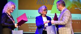 Wiceminister Machałek daje mężczyźnie w jasnej marynarce papierową torbę, obok stoi kobieta z jasnymi kręconymi włosami.