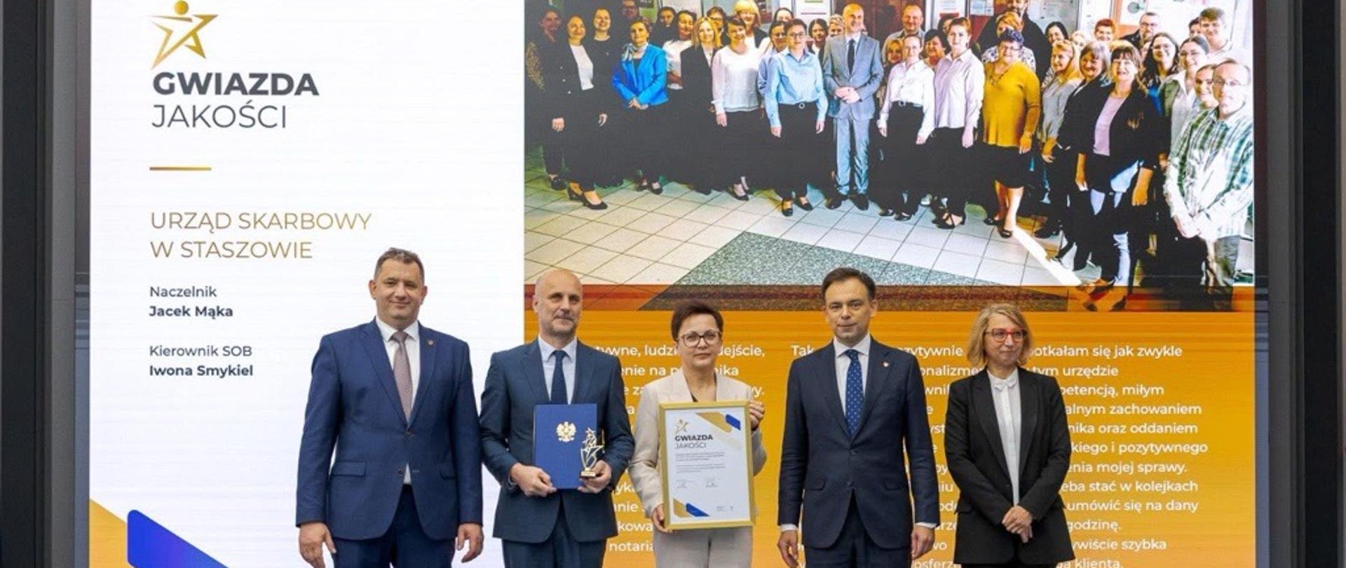 Andrzej Domański, minister finansów i gospodarki, Marcin Łoboda, szef Krajowej Administracji Skarbowej, oraz przedstawiciele Urzędu Skarbowego w Staszowie prezentują dyplom i statuetkę „Gwiazda Jakości”. Na ekranie w tle widoczna jest nazwa urzędu, zdjęcie zespołu oraz fragmenty opinii klientów.