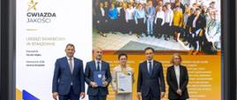 Andrzej Domański, minister finansów i gospodarki, Marcin Łoboda, szef Krajowej Administracji Skarbowej, oraz przedstawiciele Urzędu Skarbowego w Staszowie prezentują dyplom i statuetkę „Gwiazda Jakości”. Na ekranie w tle widoczna jest nazwa urzędu, zdjęcie zespołu oraz fragmenty opinii klientów
