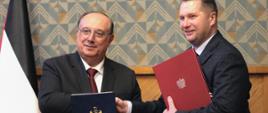 Minister Czarnek stoi obok mężczyzny w czarnym garniturze, podają sobie ręce, każdy z nich trzyma teczkę z dokumentami.
