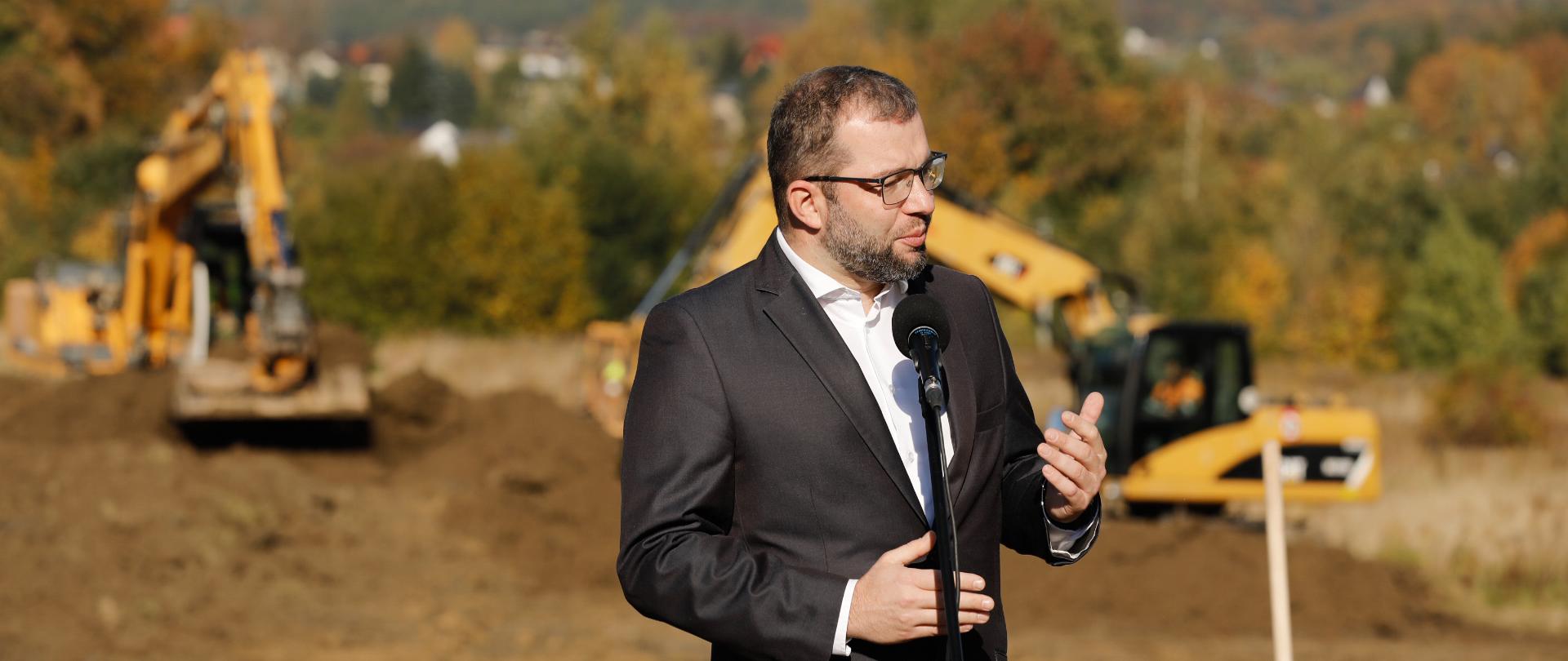 Zdjęcie przedstawia przemawiającego ministra funduszy i polityki regionalnej Grzegorza Pudę. W tle znajdują się dwie koparki.