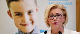 Wiceminister Machałek w białej marynarce stoi i mówi do dwóch mikrofonów, za nią ekran nas którym widać twarz usmiechniętego małego chłopca w kolorowej koszuli.