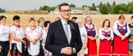 Premier Mateusz Morawiecki przy mikrofonie, za nim koło gospodyń wiejskich w strojach ludowych na tle pola.