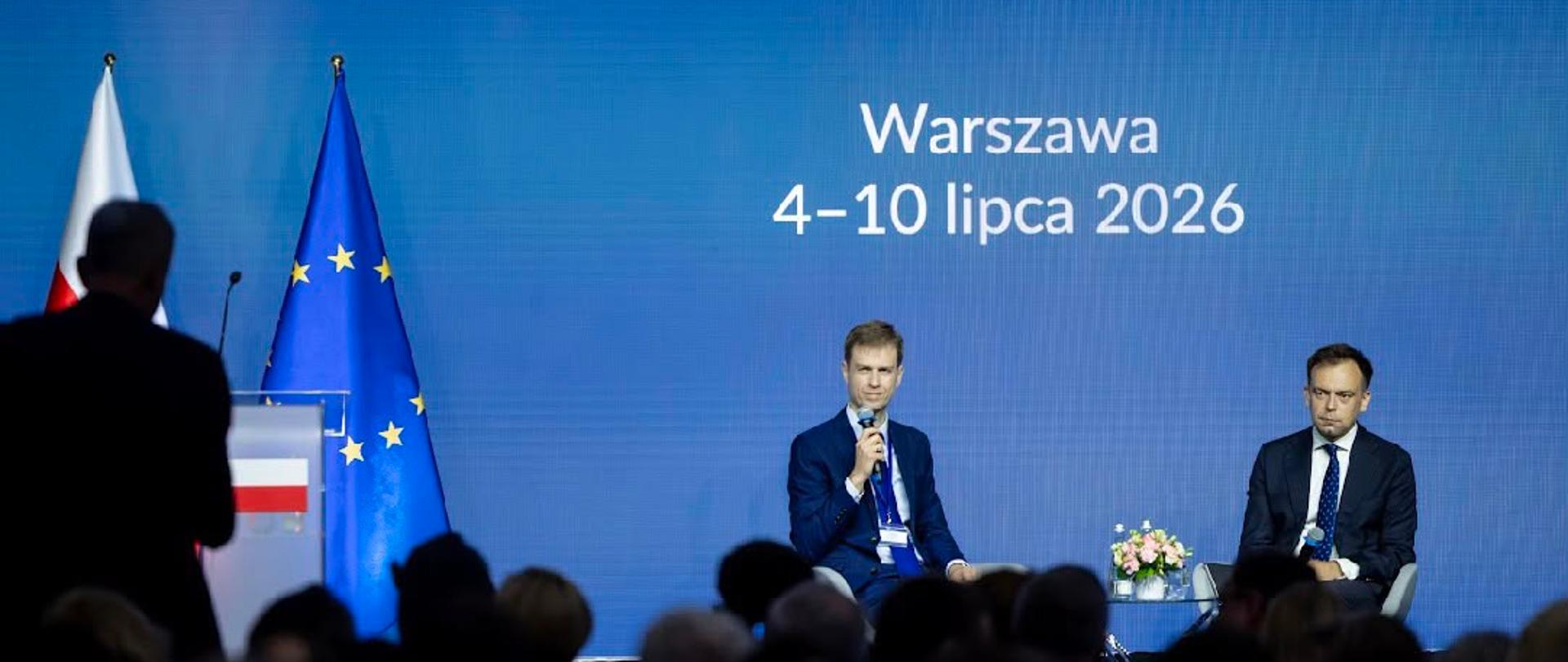 Dwie osoby uczestniczą w dyskusji na scenie podczas Narady Kierowników Placówek Zagranicznych w Warszawie, a przed sceną siedzi publiczność, za nimi widoczny jest ekran z nazwą wydarzenia oraz flagi Polski i Unii Europejskiej.