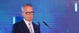 Mężczyzna w czarnym garniturze i okularach stoi przy mównicy.