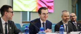 Trzy osoby siedzą z lewej strony przy stole. W środku siedzi wiceminister Jan Szyszko.