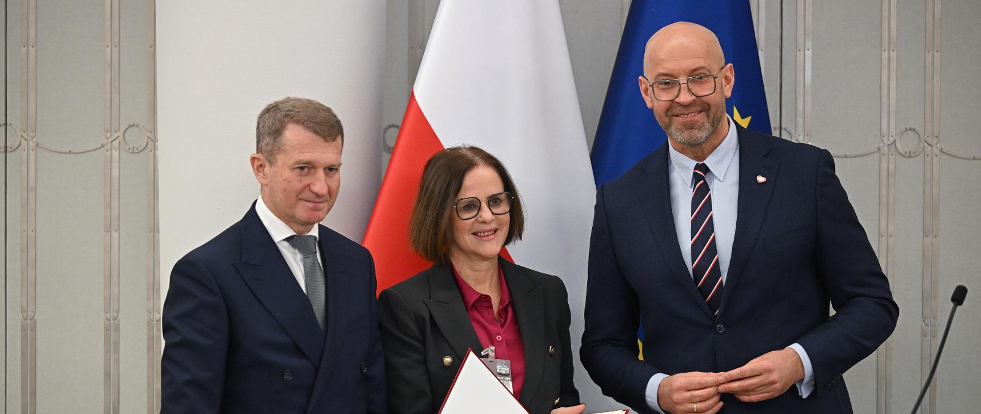 Zdjęcie. Od lewej stoją wiceminister sportu i turystyki Ireneusz Raś, nowa prezes Polskiej Organizacji Turystycznej i minister sportu i turystyki Jakub Rutnicki. Magdalena Krucz trzyma w rękach nominację na stanowisko prezesa POT. Za ich plecami flagi Polski i UE.