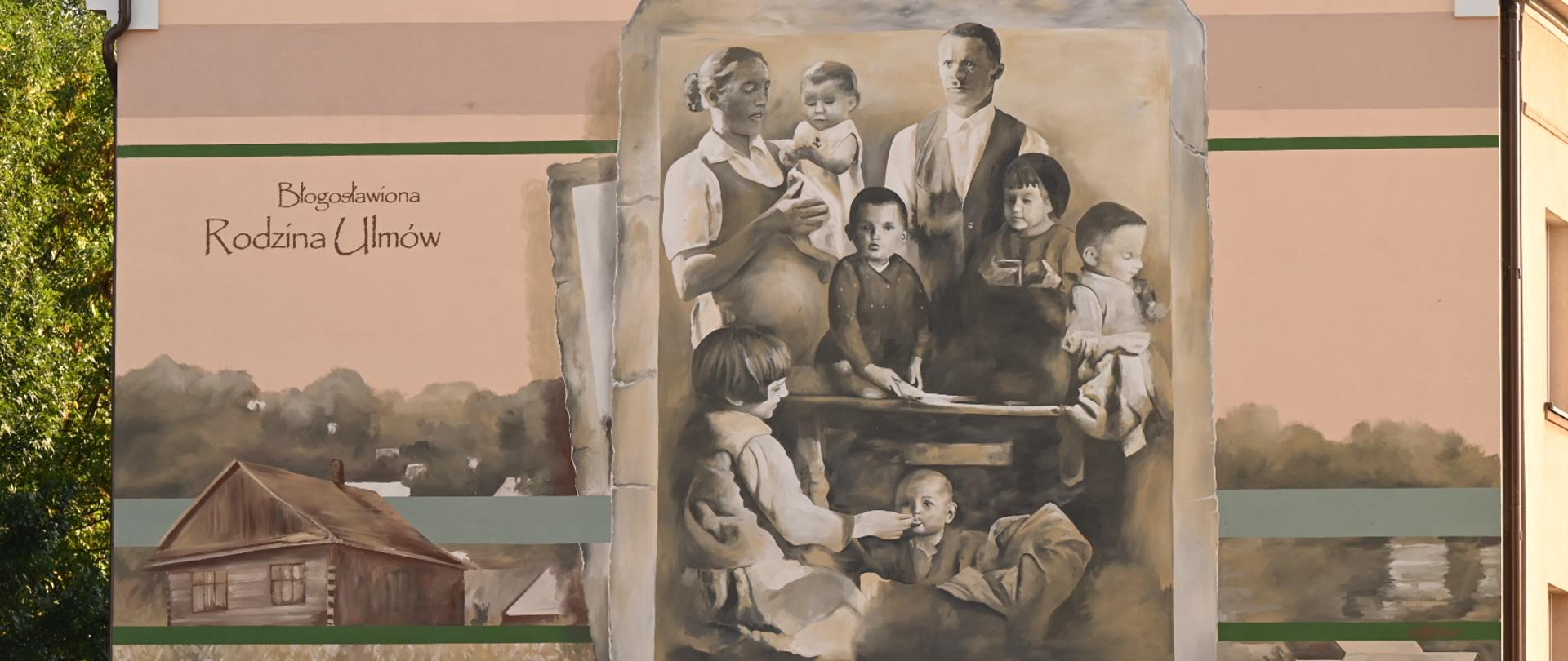 Widok na ścianę żółtego budynku, na której namalowany jest mural - odwzorowanie czarni-białego zdjęcia rodziny z sześciorgiem dzieci i napis Błogosławiona rodzina Ulmów.