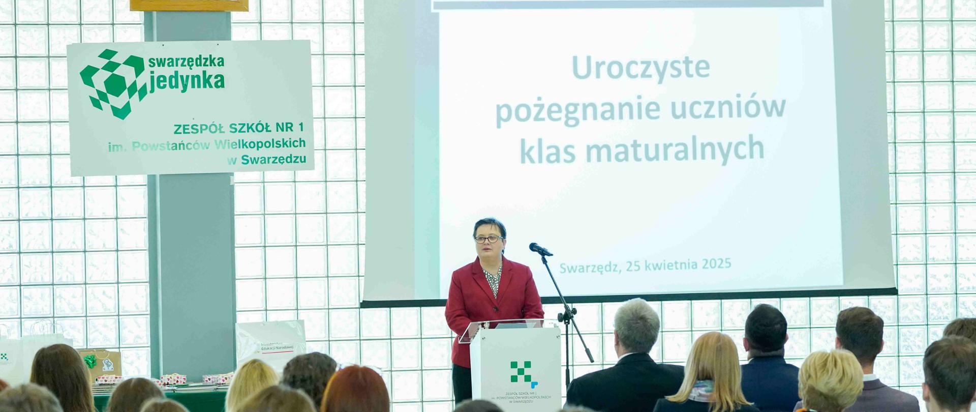 W jasnej auli, podczas uroczystości pożegnania uczniów klas maturalnych, kobieta w czerwonej marynarce stoi za mównicą i przemawia do zgromadzonej publiczności. Na mównicy widoczne jest logo szkoły. Za nią znajduje się ekran projekcyjny, na którym wyświetlany jest tytuł uroczystości: „Uroczyste pożegnanie uczniów klas maturalnych” oraz data i miejsce: „Swarzędz, 25 kwietnia 2025”.
Po lewej stronie mównicy stoi zielono-biały baner z logo szkoły „Swarzędzka Jedynka” oraz nazwą szkoły: „ZESPÓŁ SZKÓŁ NR 1 im. Powstańców Wielkopolskich w Swarzędzu”. Nad banerem wisi oprawione godło Polski.
Przed mównicą siedzi publiczność, składająca się z młodych ludzi i dorosłych. Widać ich plecy i tyły głów, co sugeruje, że są oni skupieni na przemawiającej osobie. 