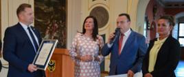 Minister stoi obok grupy osób - dwóch kolorowo ubranych kobiet i mężczyzny w garniturze, który trzyma duży symboliczny czek z napisem 5 599 200 zł.