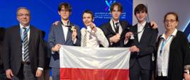 Zestawienie pięciu osób, trzech młodych mężczyzn i dwóch dorosłych, trzymających polską flagę. Młodzi mężczyźni prezentują medale, a wszyscy uśmiechają się. W tle widać fragment sceny z napisem w języku arabskim i angielskim „IChO - United Arab Emirates 2025”. Podłoga pokryta jest kolorowym konfetti.
