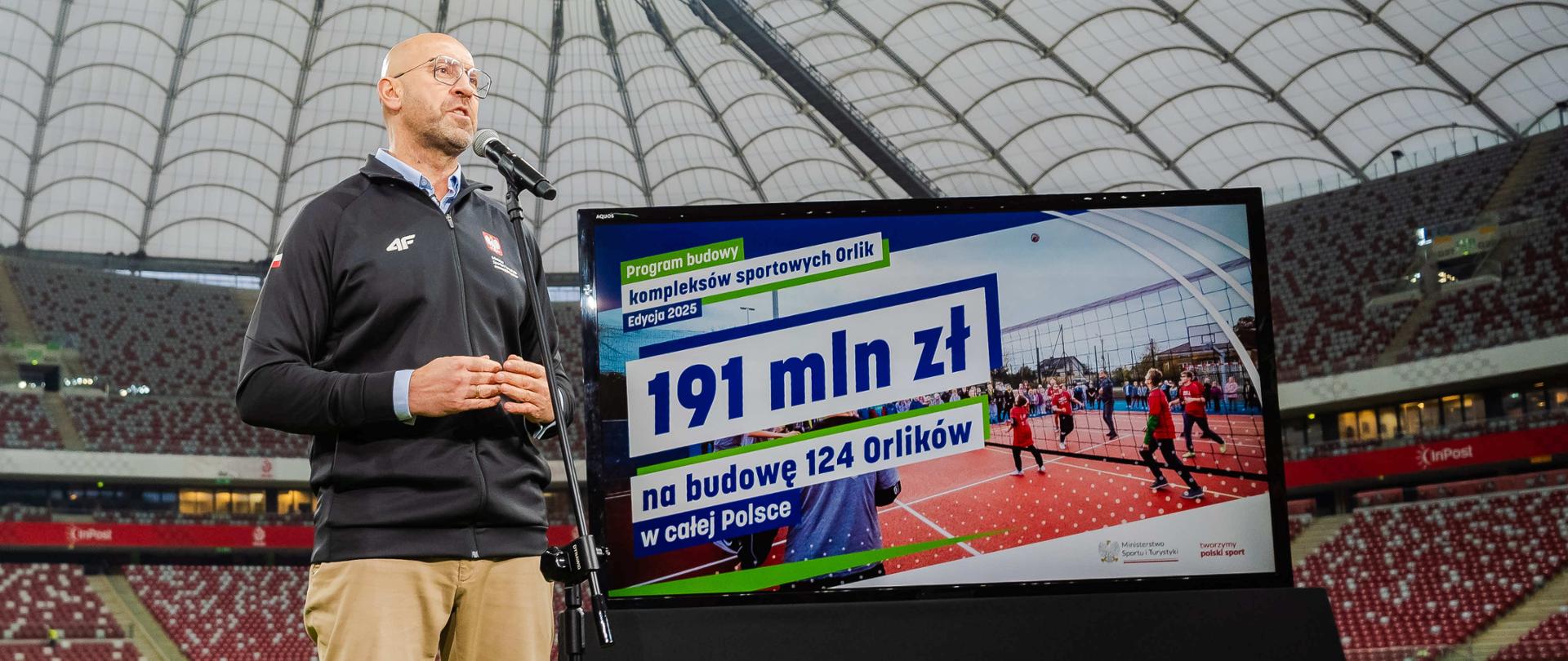 Konferencja ministra sportu i turystyki Jakuba Rutnickiego na PGE Narodowym. Minister stoi na murawie przy mikrofonie. W tle widać trybuny. Z prawej strony zdjęcia ekran z grafiką przedstawiającą podsumowanie programu budowy Orlików - edycji 2025.