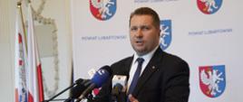 Minister Czarnek stoi za mównicą i mówi do kilku mikrofonów, za nim ścianka z niebiesko-czerwonym herbem i napisem Powiat Lubartów.