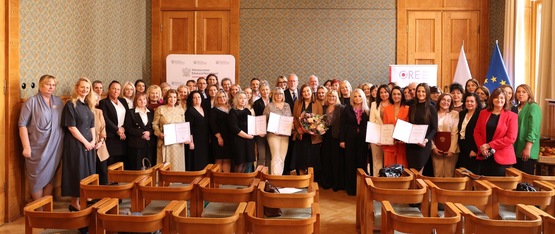 Zbiorowe zdjęcie wszystkich uczestników Gali wręczenia Krajowych Certyfikatów Szkoła i Przedszkole Promujące Zdrowie. Duża grupa osób stoi w centralnej części sali, trzymając certyfikaty i kwiaty. Nad nimi widać ozdobny, wieloramienny żyrandol.
