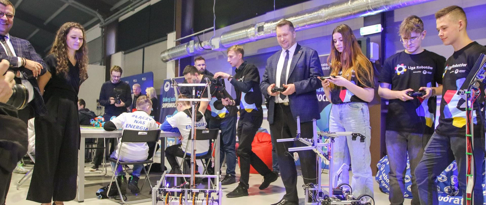 Wnętrze dużej hali, na podłodze stoi kilka robotów w postaci przestrzennych konstrukcji z metalowych kształtowników, pod ścianą stoi minister Czarnek w otoczeniu kilku osób, wszyscy mają w rękach piloty zdalnego sterowania.