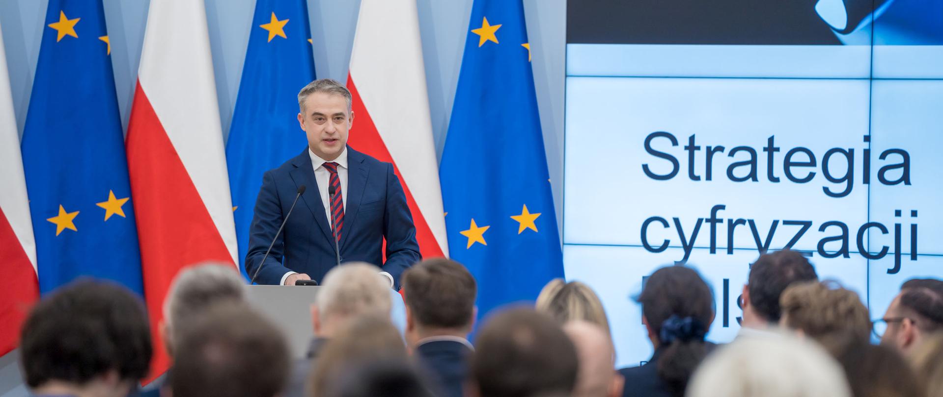 Na zdjęciu widzimy wicepremiera i ministra cyfryzacji Krzysztofa Gawkowskiego, który wygłasza przemówienie podczas wydarzenia „Strategia Cyfryzacji do 2035 roku”. Za ministrem znajdują się flaga Polski oraz flaga Unii Europejskiej. Po prawej stronie ministra widać fragment prezentacji z napisem „Strategia Cyfryzacji”. Przed ministrem widoczna jest publiczność.