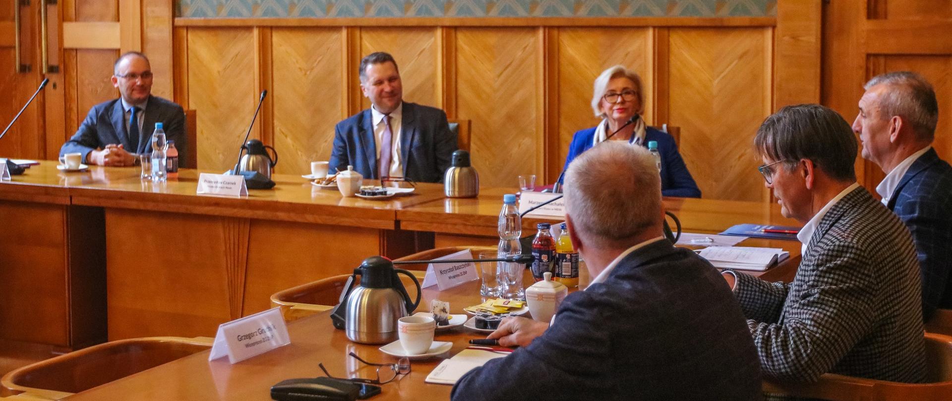 Kilka osób, wśród nich minister Czarnek i wiceminister Machałek, siedzi przy stole i rozmawia.