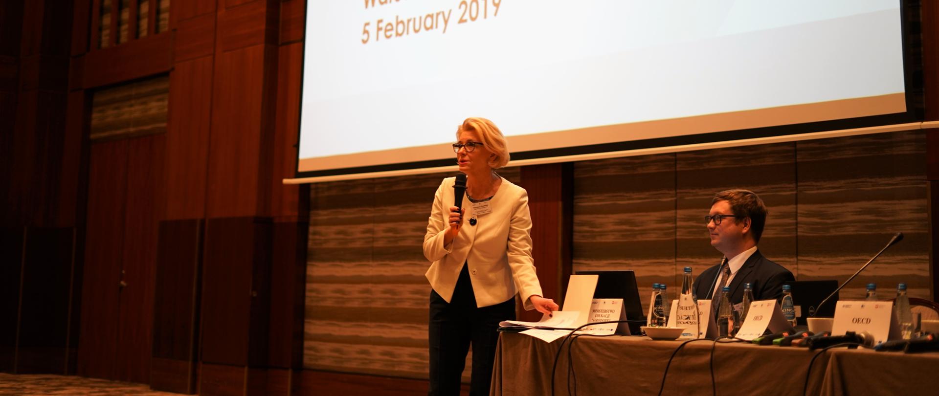 wiceminister Marzena Machałek przemawia do uczestników, w tle prezentacja