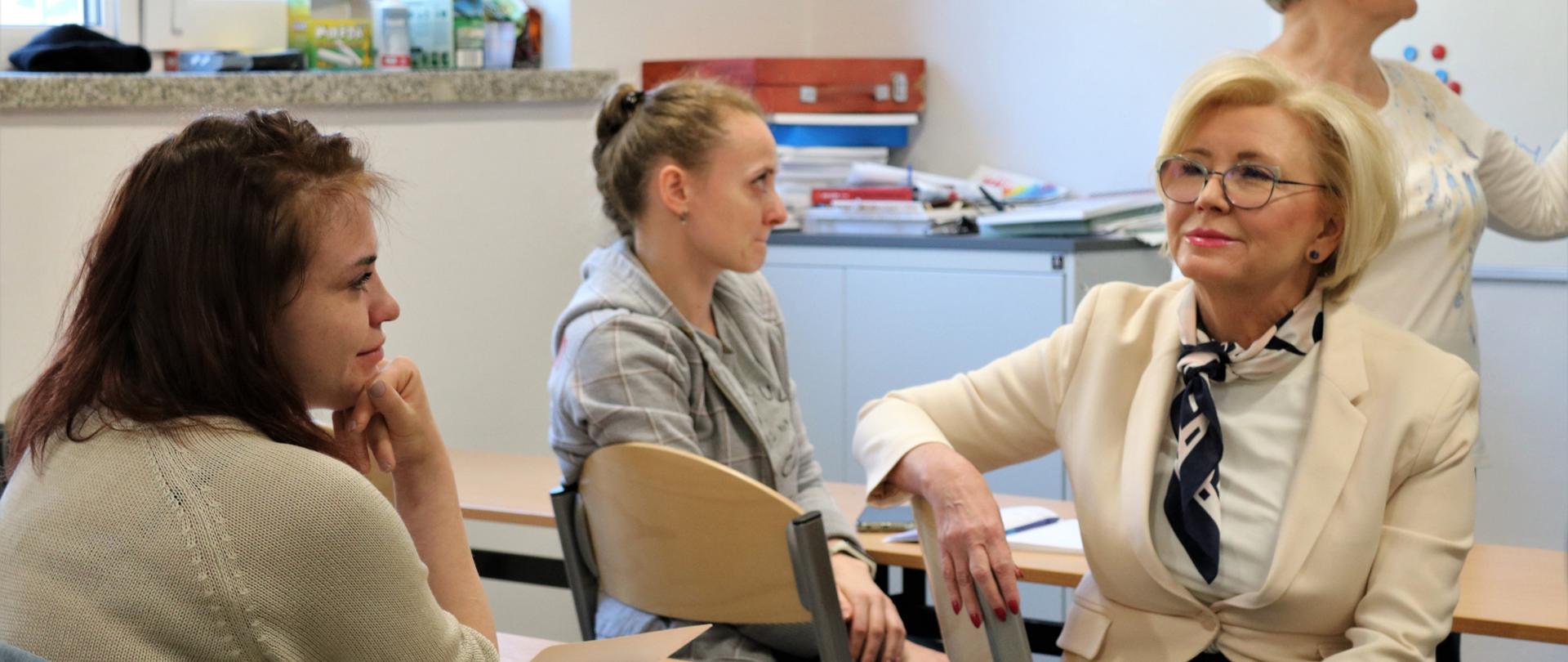 Dwie dziewczynki siedzą w ławkach szkolnych. Naprzeciw nich wiceminister Marzena Machałek ubrana w biały żakiet, Ma okulary. W tle gestykulująca osoba. 