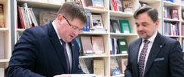 Wiceminister Rzymkowski ogląda leżącą na okrągłym białym stoliku grubą książkę w kolorowej okładce, obok niego stoi mężczyzna w garniturze, za nimi półki z książkami.
