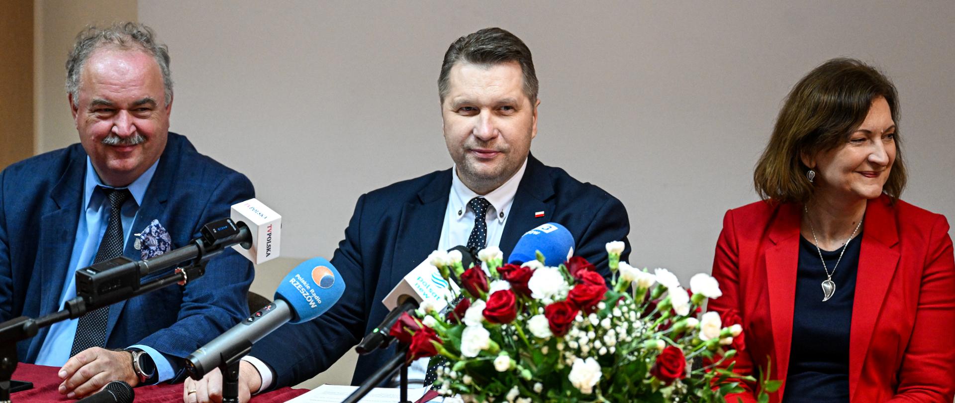 Dwóch siedzących mężczyzn przy mikrofonach i kobieta, przed nimi kwiaty