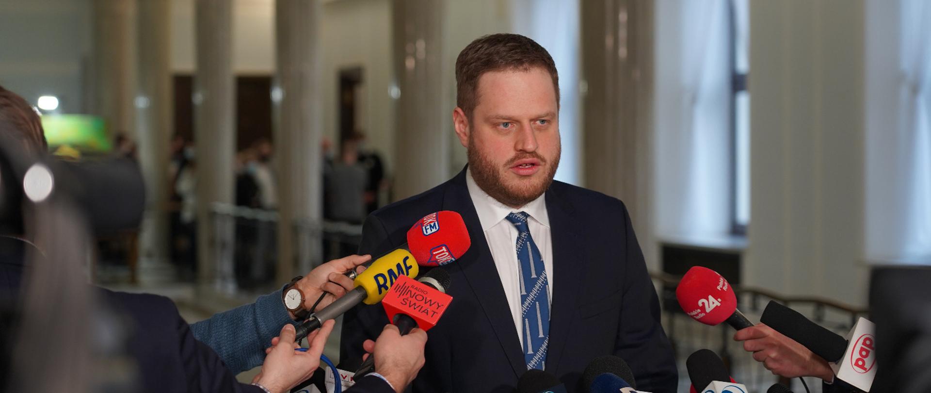 Na głównym planie Minister Jan Cieszyński udzielający wywiadu. Przed nim wyciągnięte ręce trzymające mikrofony. 