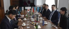 Zdjęcie z boku, przy drewnianym stole siedzi grupa elegancko ubranych osób, przed nimi na stole małe flagi Polski i Japonii, za nimi białe ściany, w nich drewniane drzwi przed którymi stoi duży wyłączony monitor, z prawej zasłonięte białą firanką okno.