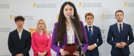Młoda dziewczyna w fioletowym sweterku stoi przy mikrofonie na stojaku, za nią trzech młodych mężczyzn w garniturach i dziewczyna w czerwonej sukience, za nimi biała ściana z napisem Rada Dzieci i Młodzieży.