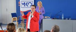 Wiceminister Katarzyna Lubnauer i Mazowiecka Kurator Oświaty stoją na scenie i przemawiają do siedzących na auli osób. Wiceminister ma w ręku mikrofon i zabiera głos. Mazowiecka Kurator Oświaty stoi za nią i przysłuchuje się. Zdjęcie wykonane jest zza pleców siedzących na auli uczestników spotkania. 