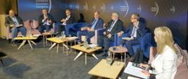 Widok z boku na salę, pod czarną ścianą z napisem European Economic Congress na fotelach za małymi stolikami siedzi kilka osób, pośrodku wiceminister Murdzek mówi do trzymanego w ręku mikrofonu.