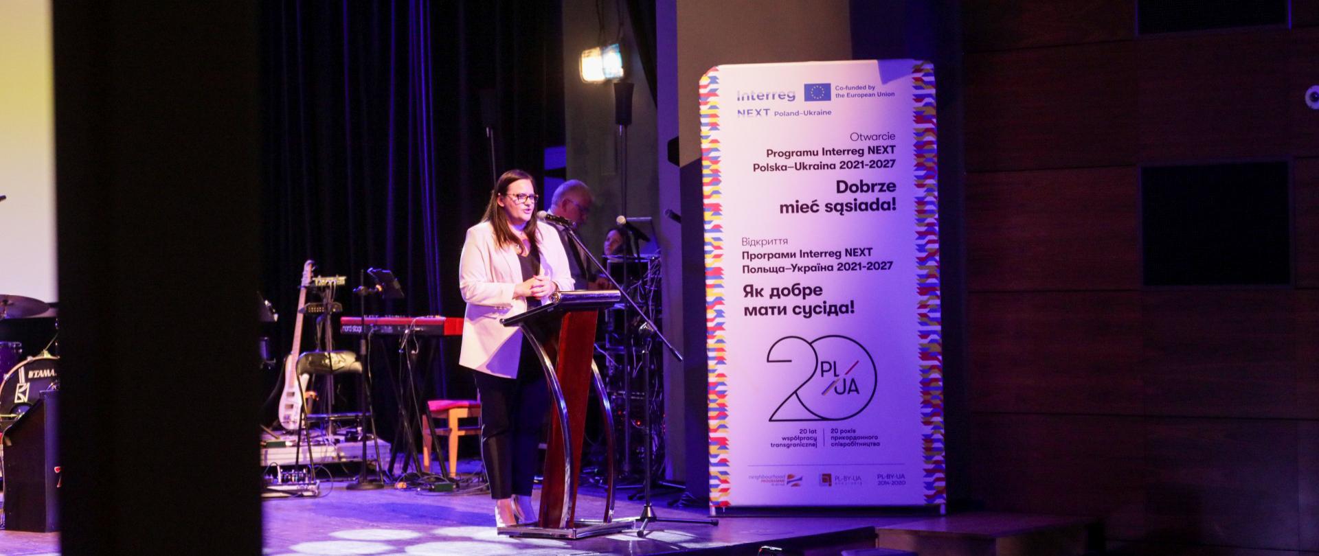 Wiceminister Małgorzata Jarosińska-Jedynak stoi na scenie przy pulpicie i mówi do widzów.