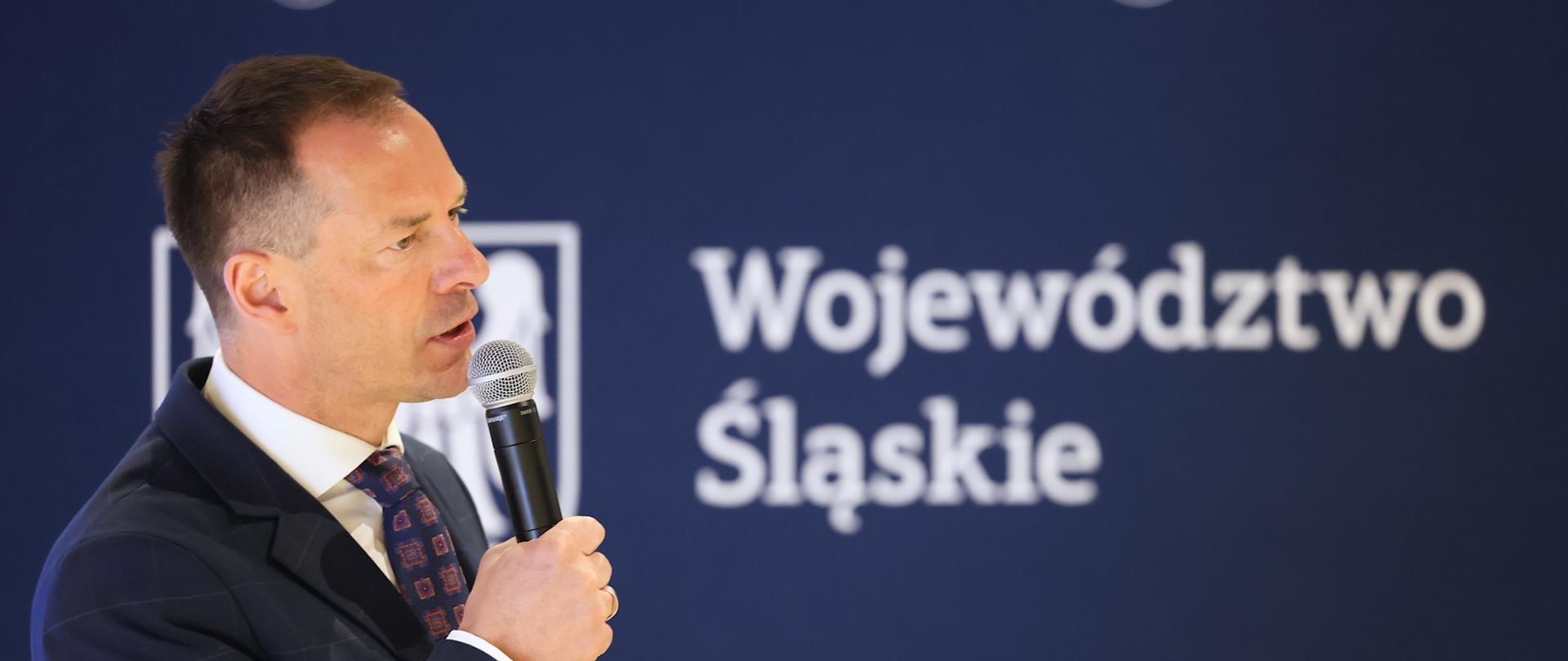 Piotr Borys na konferencji w Szczyrku. Minister trzyma w ręku mikrofon, za nim stoi ścianka z logiem województwa śląskiego