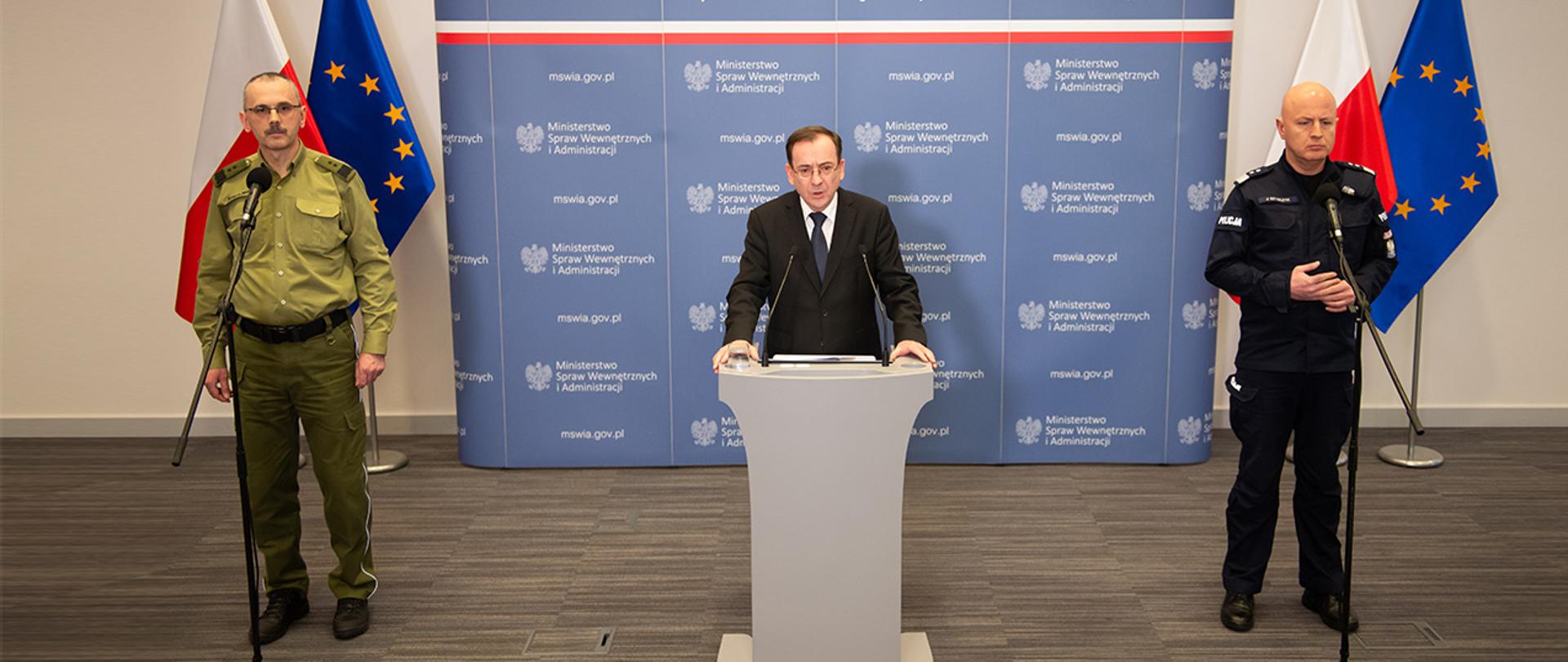 Na zdjęciu widać Mariusza Kamińskiego ministra SWiA stojącego za mównicą i przemawiającego. Widać również gen. dyw. SG Tomasza Pragę komendanta głównego Straży Granicznej i gen. insp. Jarosława Szymczyka komendanta głównego Policji. W tle stoi scianka MSWiA i flagi Polski i UE. 