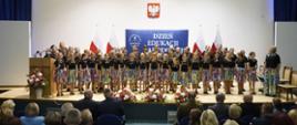 Zdjęcie od tyłu sali, na podwyższeniu z przodu stoi duża grupa dziewczynek w czarnych koszulkach i kolorowych spódniczkach, z boku stoi kobieta w czarnej koszulce, przed nimi na podłodze leżą bukiety biało-czerwonych kwiatów.