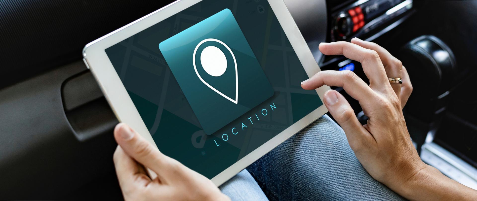 Kobieta siedząca w samochodzie i obsługująca tablet z nawigacją. Na ekranie tabletu symbol wskaźnika miejsca oraz napis "Location".