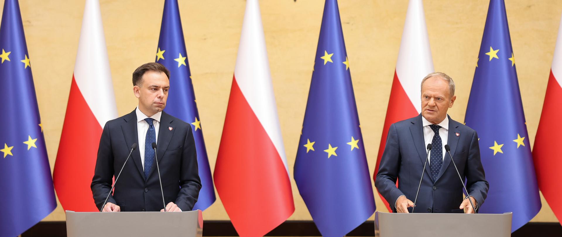 Dwie osoby stoją przy mównicach na tle flag Polski i Unii Europejskiej. Nad nimi widoczny jest godło Rzeczypospolitej Polskiej.