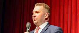 Minister Czarnek stoi i mówi do trzymanego w ręku mikrofonu, za nim czerwona zasłona.